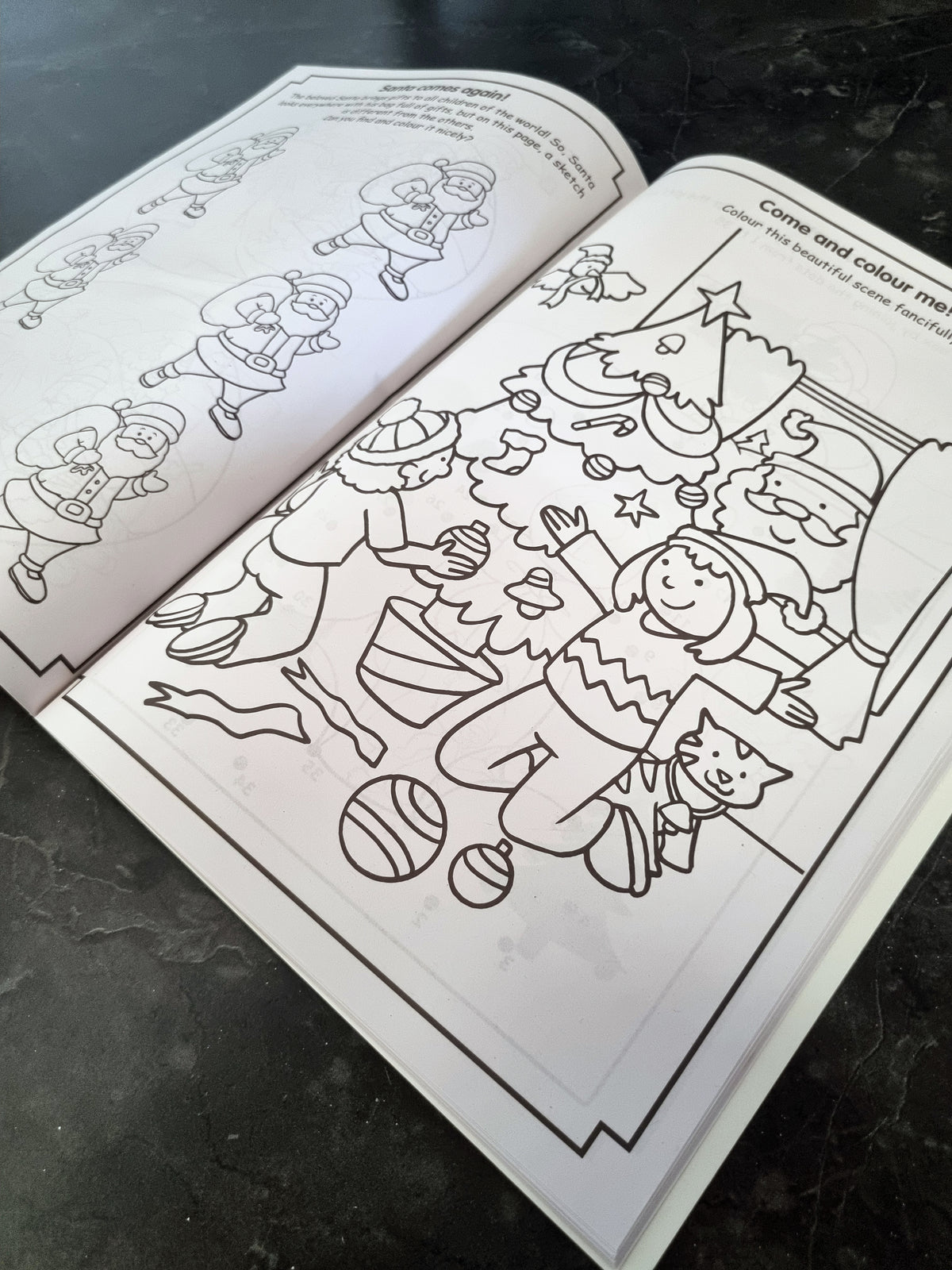 An open children's coloring book showing a black-and-white Christmas scene with a child, tree, ornaments, and Santa, photographed on a dark countertop.