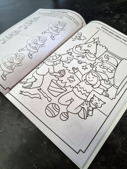 An open children's coloring book showing a black-and-white Christmas scene with a child, tree, ornaments, and Santa, photographed on a dark countertop.