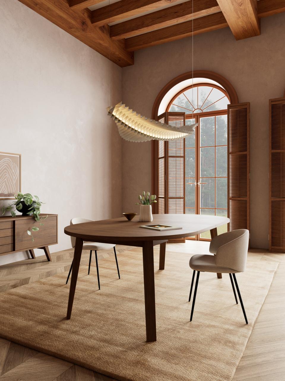 A modern dining room scene featuring a round wooden dining table with dark wood legs, surrounded by neutral upholstered chairs, set on a large beige rug under a hanging woven pendant light. The room has warm plaster walls, exposed wooden ceiling beams, and an arched window with shutters.