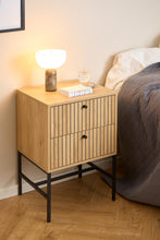 Load image into Gallery viewer, Wooden nightstand with lamp and glass on a wooden floor next to a bed.
