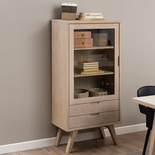 Load image into Gallery viewer, A tall light oak wood cabinet with a glass-front upper door, two lower drawers, and angled legs, styled with books and baskets against a neutral beige wall.
