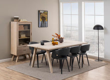 Load image into Gallery viewer, A modern dining room setup featuring a rectangular light wood table with angled legs, surrounded by six black chairs. The table is styled with a vase of orange flowers and two amber glasses, set on a textured gray rug.
