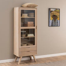 Load image into Gallery viewer, A tall light wood display cabinet with glass doors and drawers stands against a beige wall on a wood floor, styled with books, baskets, and decorative boxes. A framed painting and a woven pillow are visible in the room setting.
