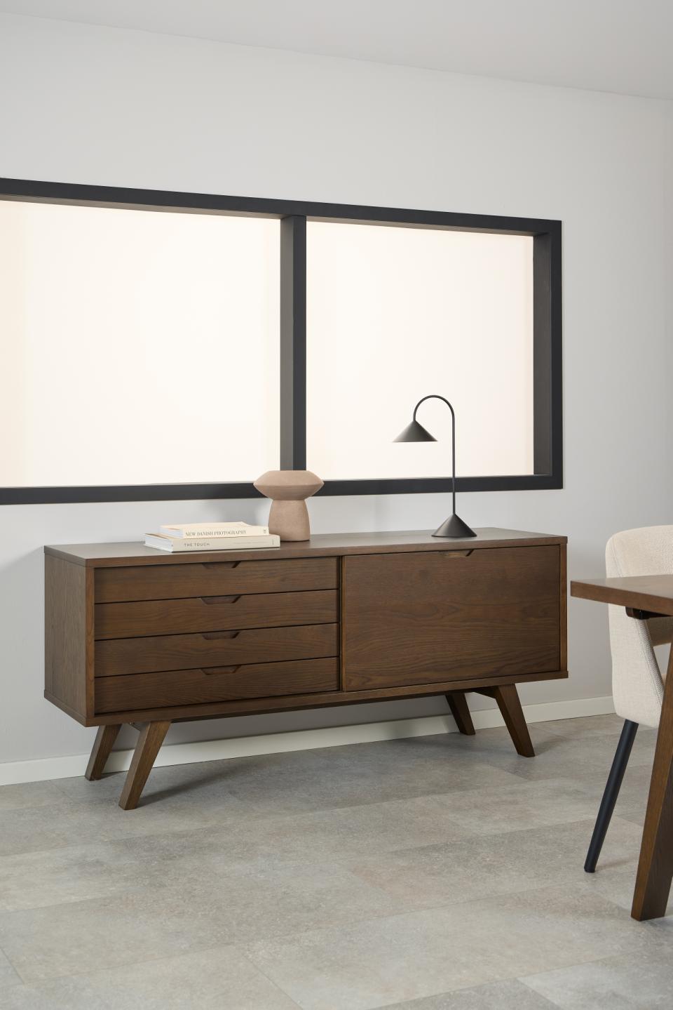 A mid-century modern wooden sideboard or credenza in a warm brown finish, styled in a minimalist room with a black-framed window, a black floor lamp, and a light gray tile floor.
