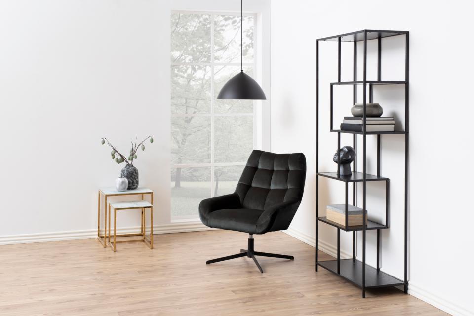 A modern black leather swivel armchair in a minimalist room with light wood floors, a black pendant light, a small gold side table, and a tall black metal shelving unit.