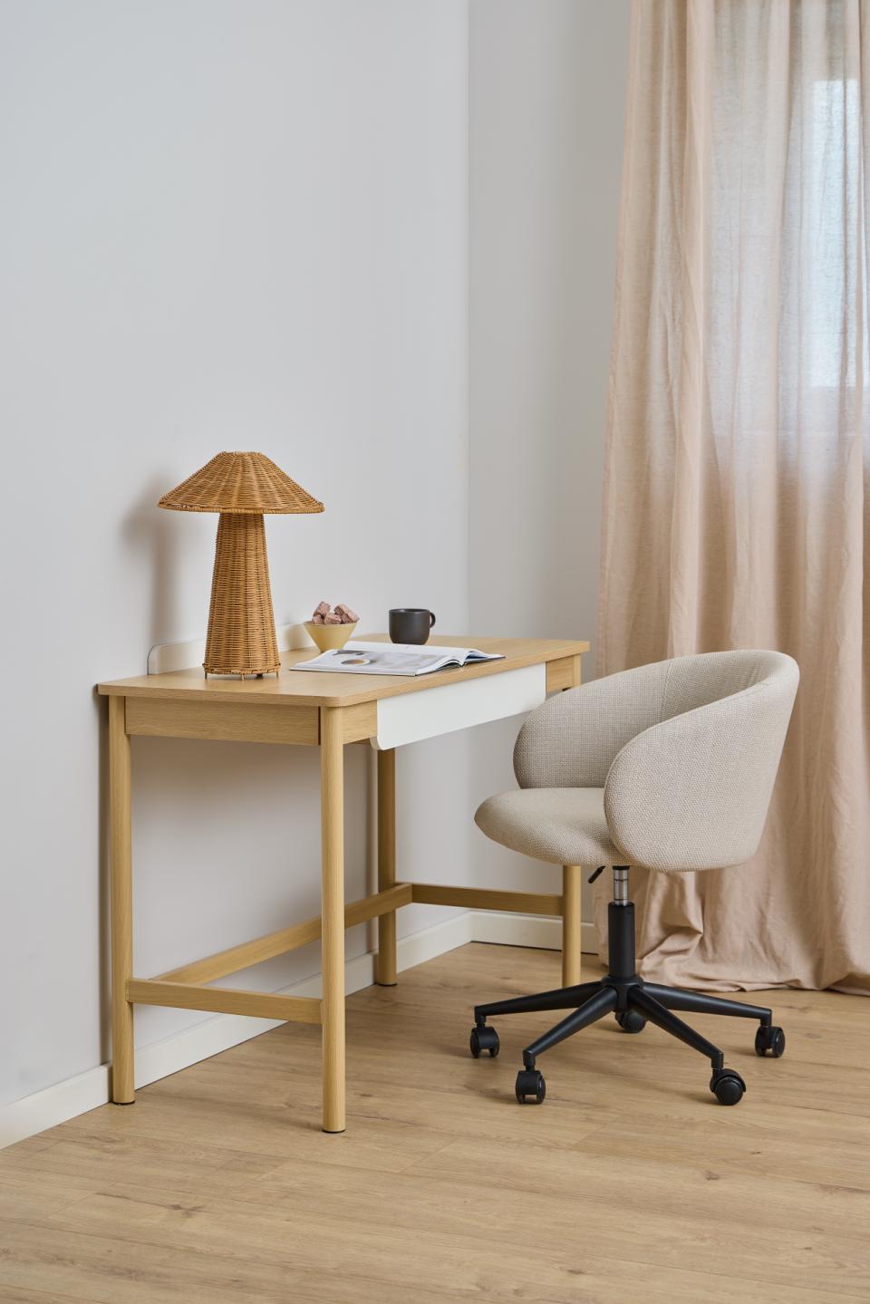 A light wood writing desk with a white drawer front and a beige upholstered swivel chair, styled in a bright room with a wicker lamp, curtains, and a light wood floor.