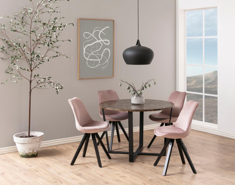 A round dining table with a dark stone-look top and black metal base, surrounded by four blush pink upholstered chairs with black legs. The dining set is staged in a modern dining room with light wood floors, a gray wall, a black pendant light, and a potted tree.