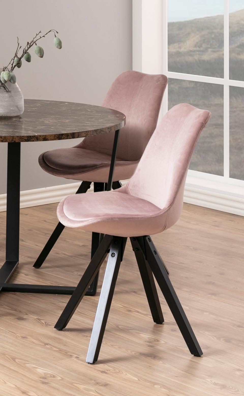 A modern dining chair in light pink velvet with black splayed legs, positioned in a bright room next to a round dark wood table. A second matching chair in a dusty mauve-brown velvet is visible in the background near a large window.