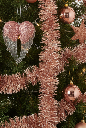 Rose gold Christmas tree ornaments including a glittery heart-shaped angel wings ornament, shiny metallic baubles, and tinsel garland hanging on a green Christmas tree.
