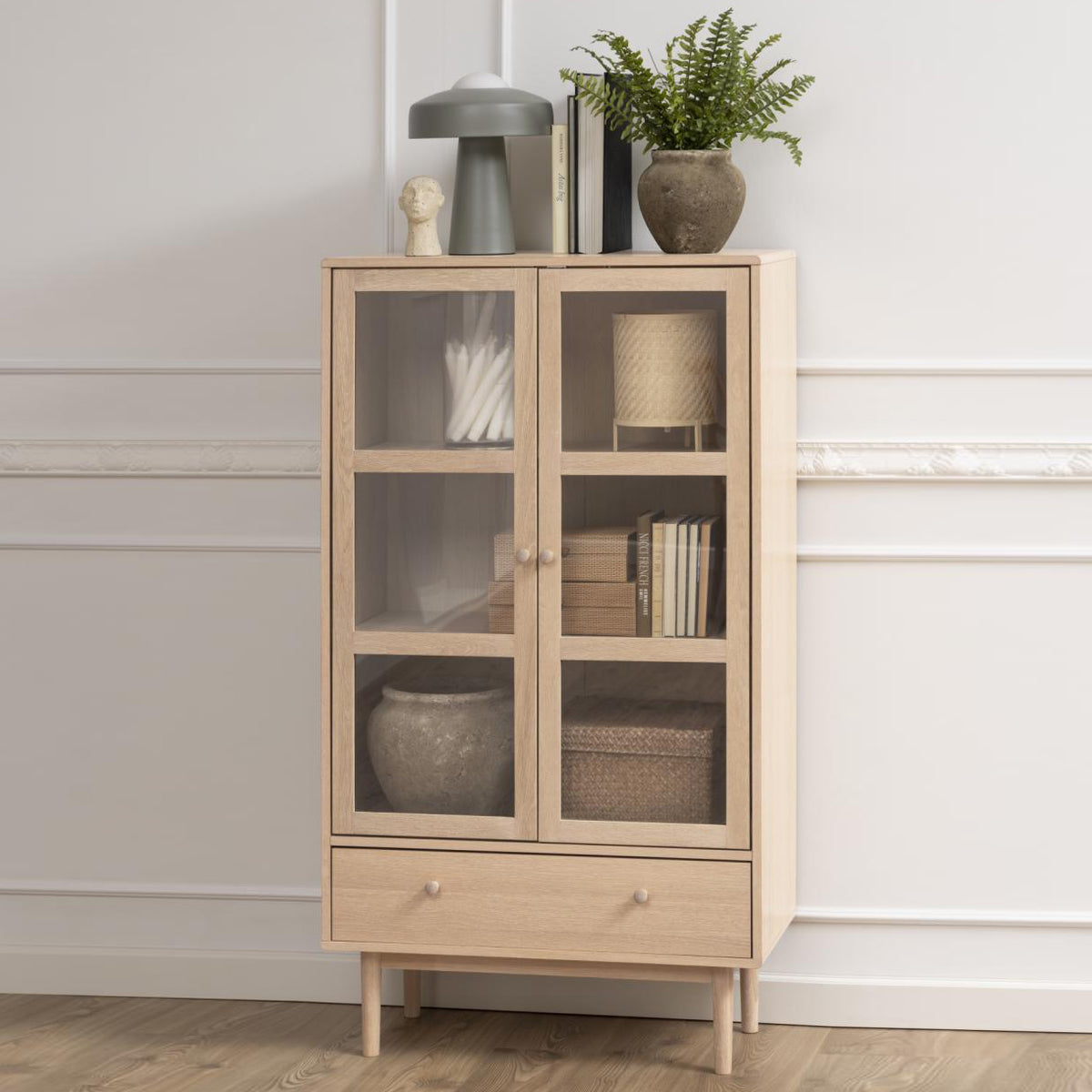 A white oak display cabinet with two glass doors, shelves, and a drawer. The cabinet is positioned in a room with decorative items displayed on top and inside.