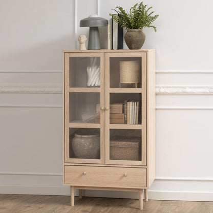 A white oak display cabinet with two glass doors, shelves, and a drawer. The cabinet is positioned in a room with decorative items displayed on top and inside.