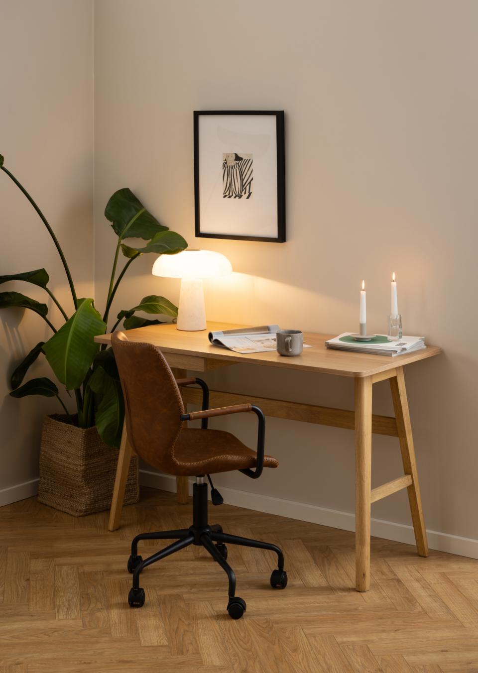 A warm, modern home office corner featuring a light wood desk with a brown leather swivel chair, styled with a lit table lamp, candles, and a framed print on the beige walls.