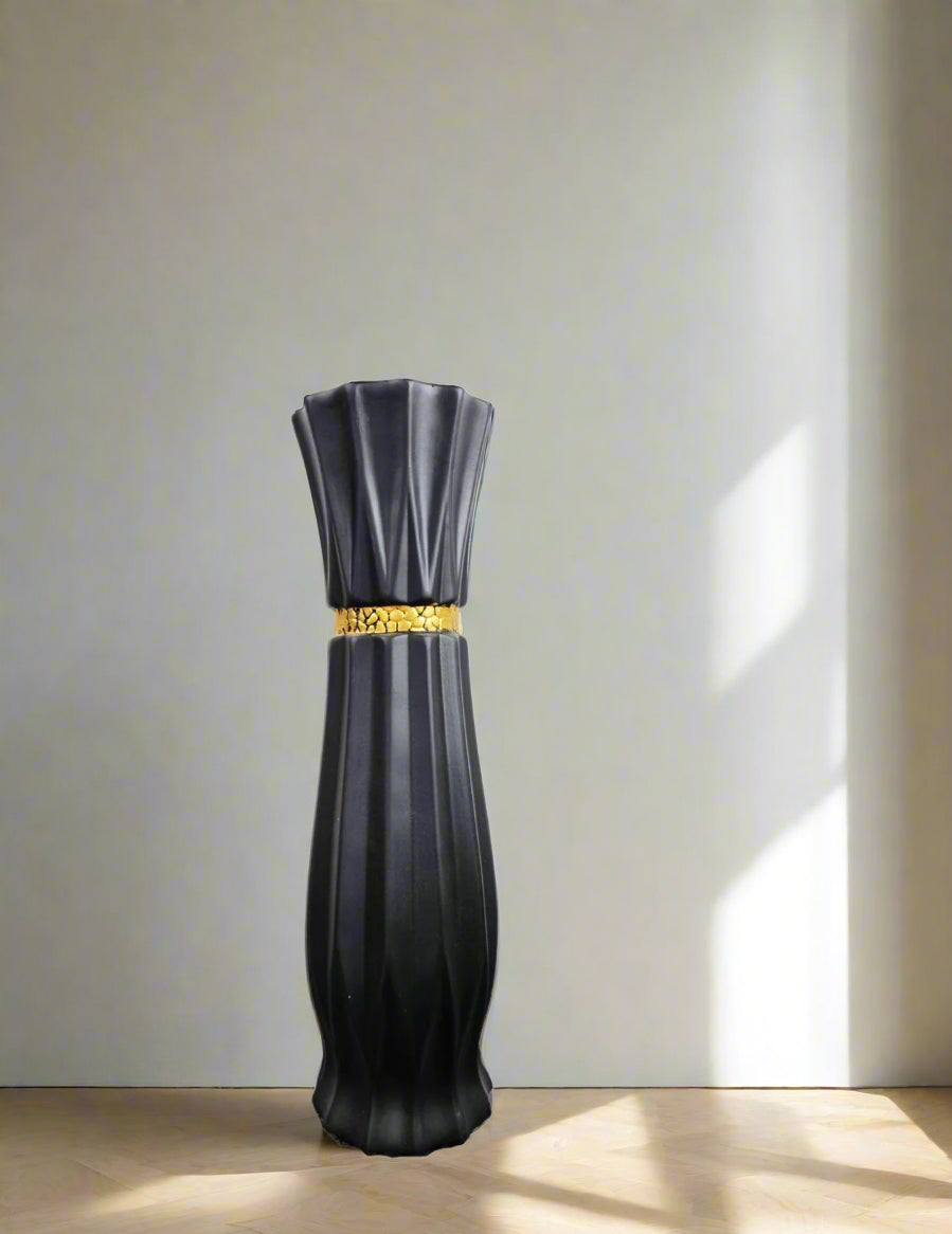 A tall sculptural black vase with a flared top and a textured gold band around the middle, standing on a light wood floor against a warm beige wall with soft sunlight streaming in from the side.