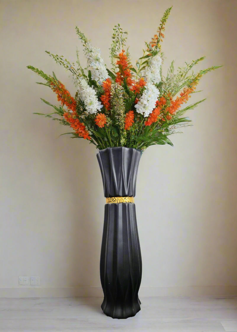 A tall black fluted vase with a gold band around the neck, filled with a large floral arrangement of orange, white, and green flowers, set against a plain beige wall.