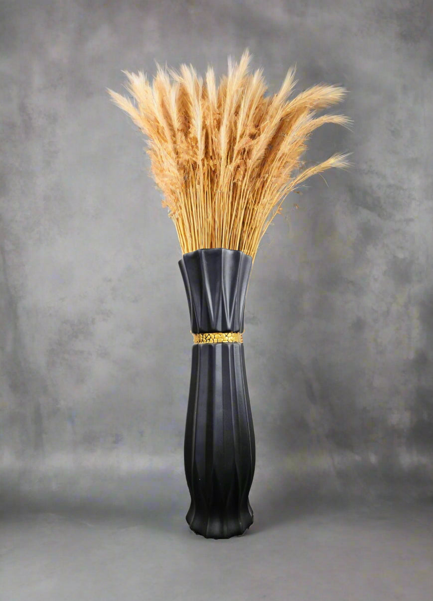 A black ribbed ceramic vase with a gold band detail, filled with dried golden wheat stalks, set against a textured gray background.