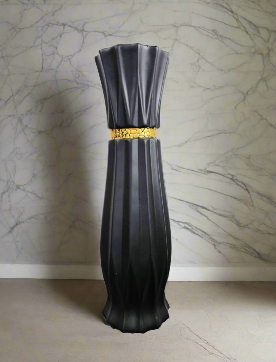 A tall, fluted black ceramic vase with a textured gold band around the middle, set against a gray marble-patterned wall.