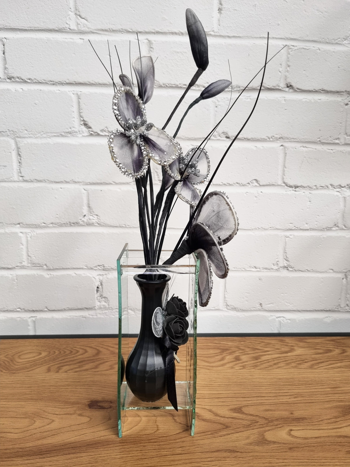 A decorative floral arrangement in a clear rectangular glass vase with a black bulb-shaped vase and silver-gray artificial flowers, set on a wooden surface against a white brick wall.