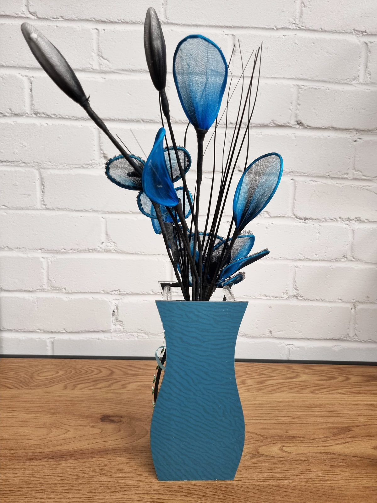 A decorative arrangement of blue and black artificial flower stems in a textured blue vase, set on a wooden surface against a white brick wall.