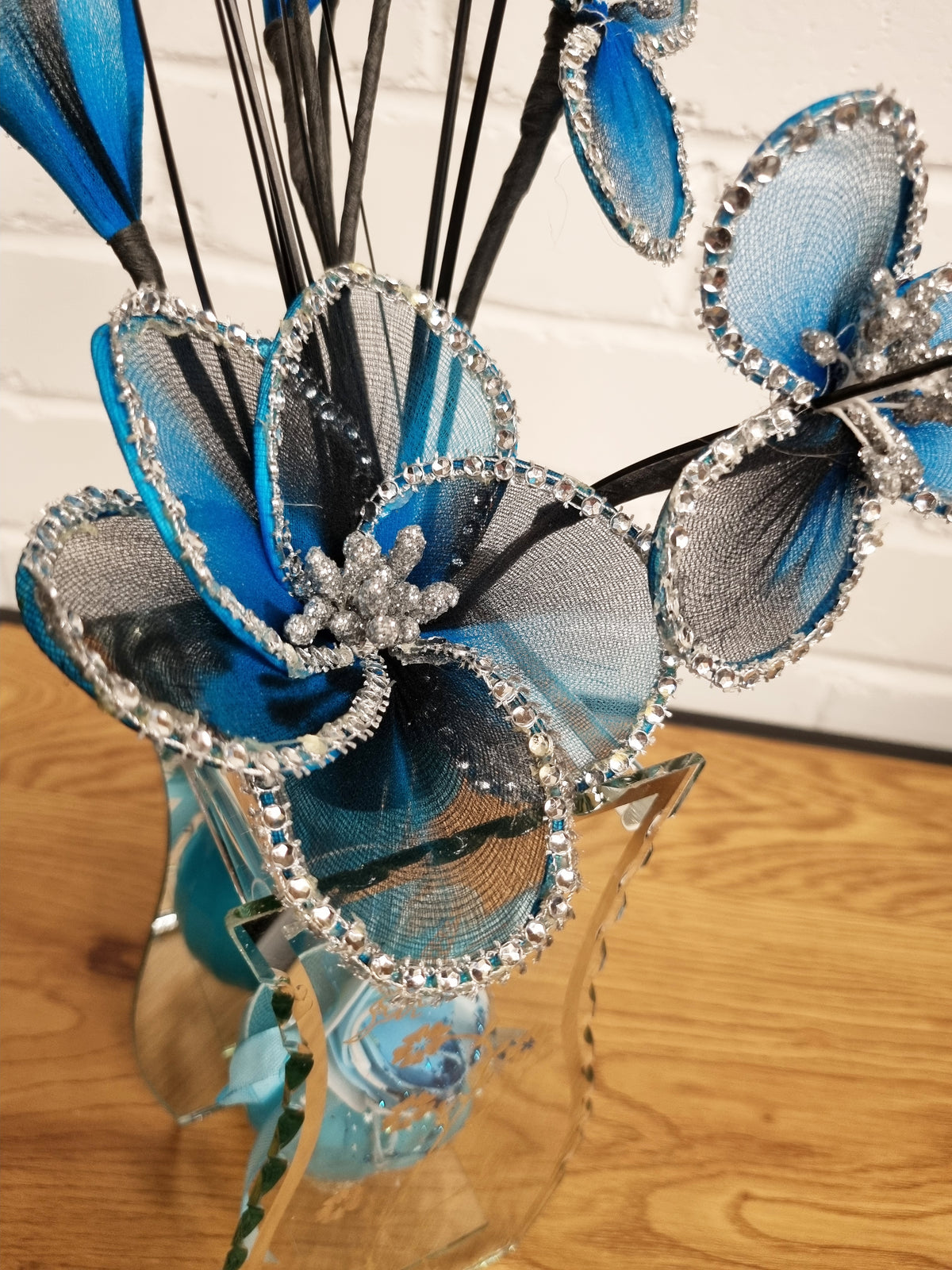 Blue and black mesh flower hair sticks with silver rhinestone trim, arranged in a clear glass vase on a wooden surface against a white brick wall.