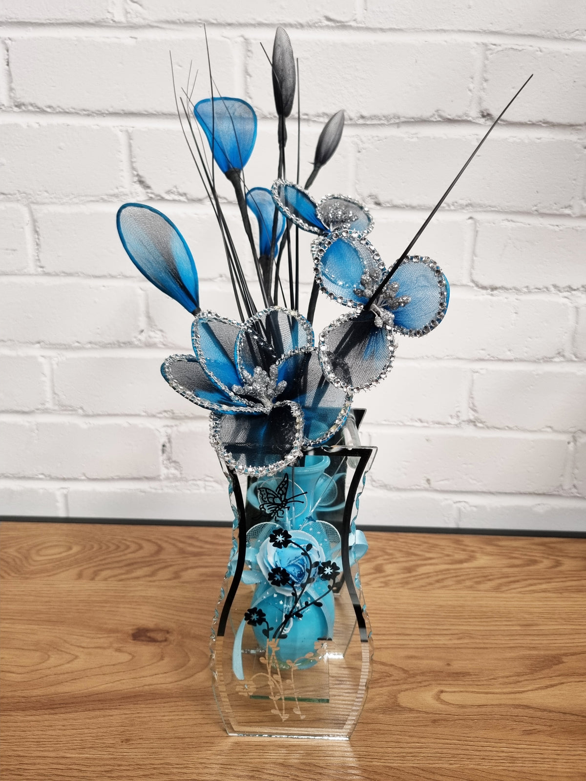 A decorative floral arrangement in a clear glass vase, featuring blue and black artificial flowers, butterfly-shaped elements, and feather-like stems, set on a wooden surface against a white brick wall.
