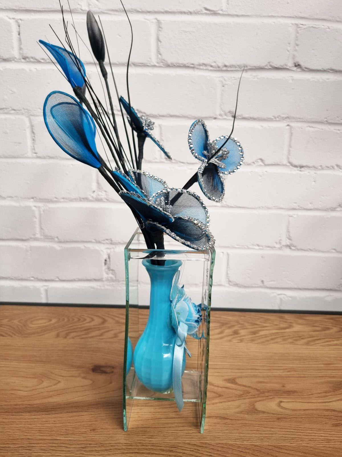 A decorative floral arrangement featuring blue and black artificial flowers with silver glitter accents, displayed in a clear glass vase with a turquoise blue neck, set on a wooden surface against a white brick wall background.