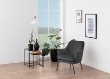 Load image into Gallery viewer, A modern gray upholstered armchair in a bright, minimalist room with light wood floors, a large window, potted plants, a black side table, and framed wall art.
