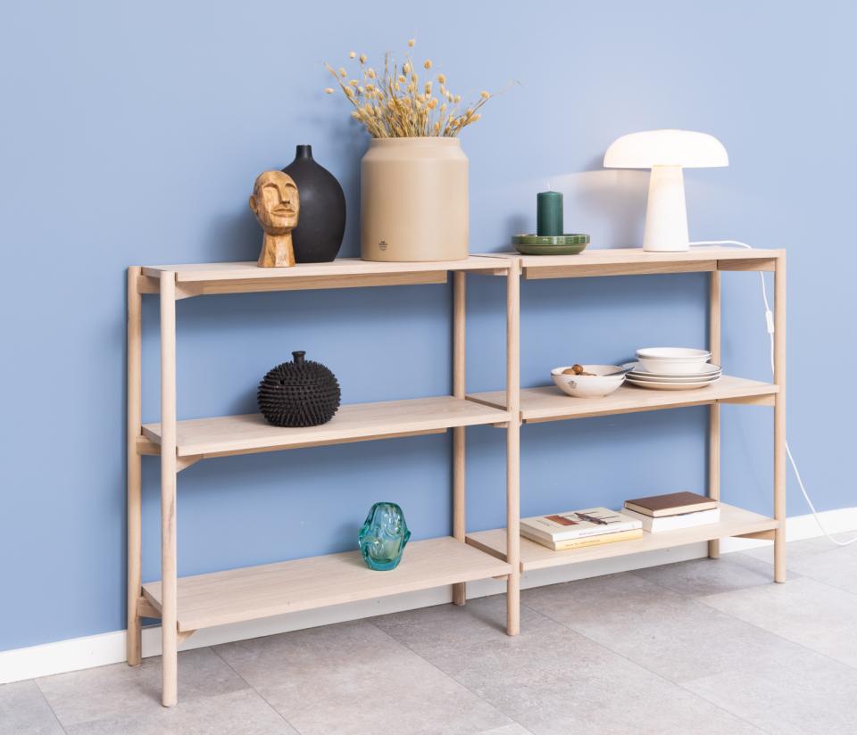 A light natural wood four-tier shelving unit styled with home decor items against a blue wall and gray floor.