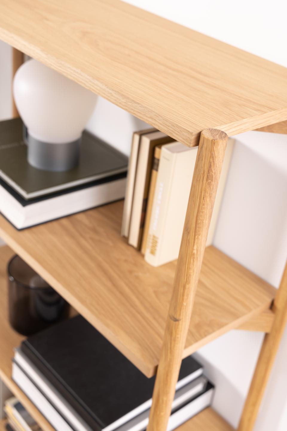 Close-up of a light natural wood bookshelf with open shelves, styled with books and a white globe lamp against a white wall.
