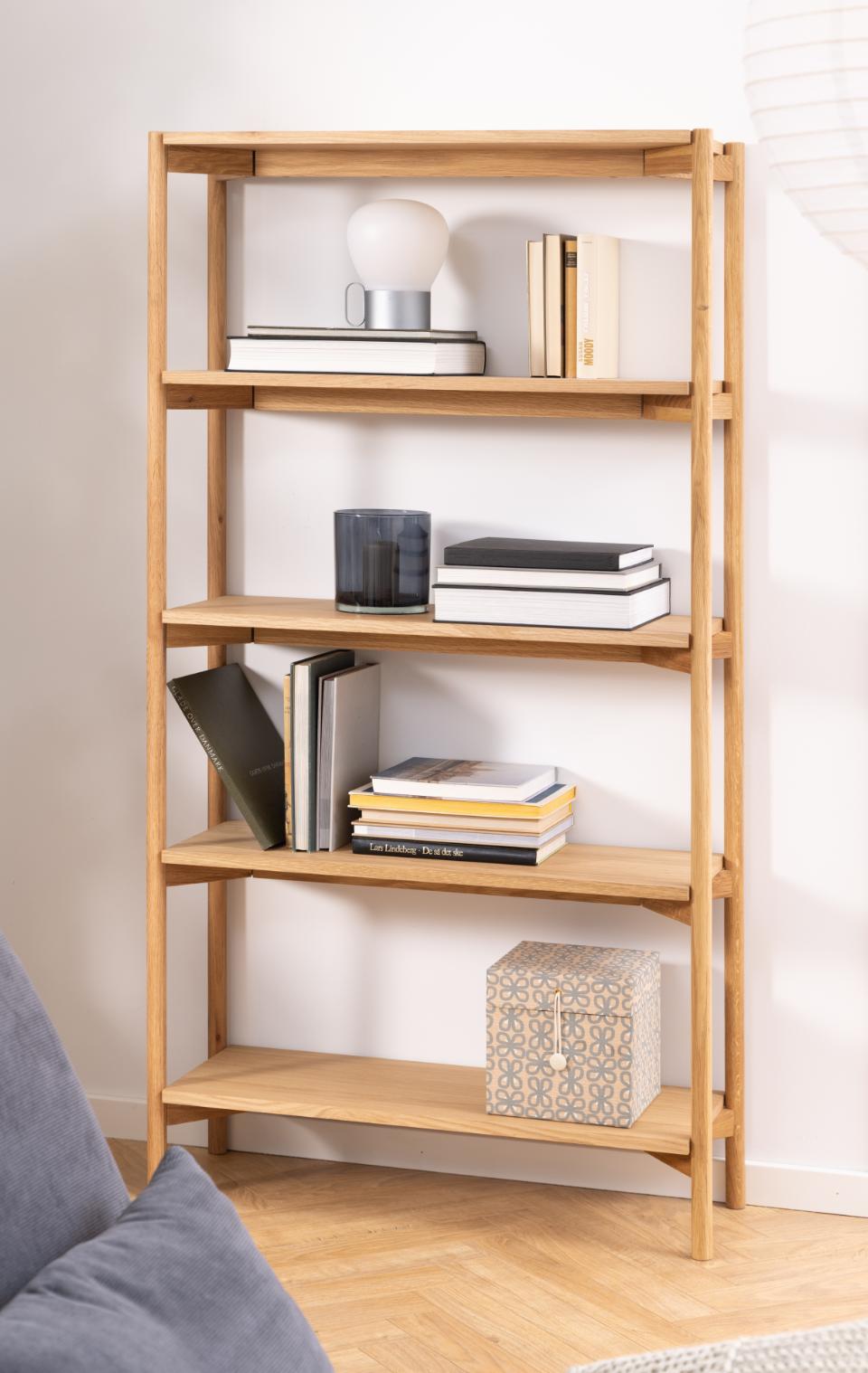 A light natural wood 5-tier bookshelf styled with books, a lamp, and decorative boxes in a bright room.