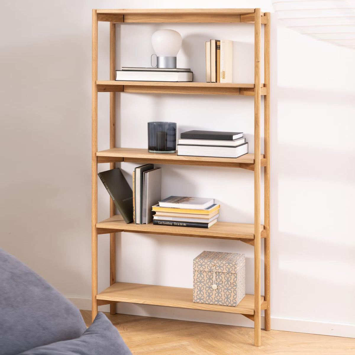 A tall, five-tier wooden bookshelf with a natural light oak finish, styled with books, a lamp, and decorative boxes in a bright room.