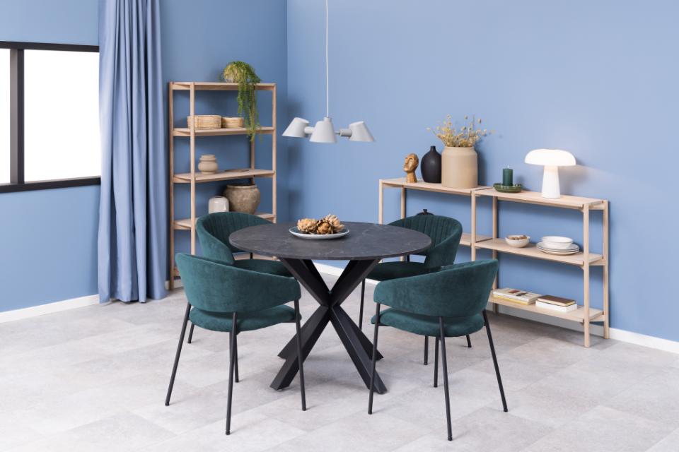 A modern dining room setup featuring a round black table with a black X-shaped base, surrounded by four dark teal velvet chairs. The room has light blue walls, a light gray tiled floor, and light wood shelving units decorated with home accessories.
