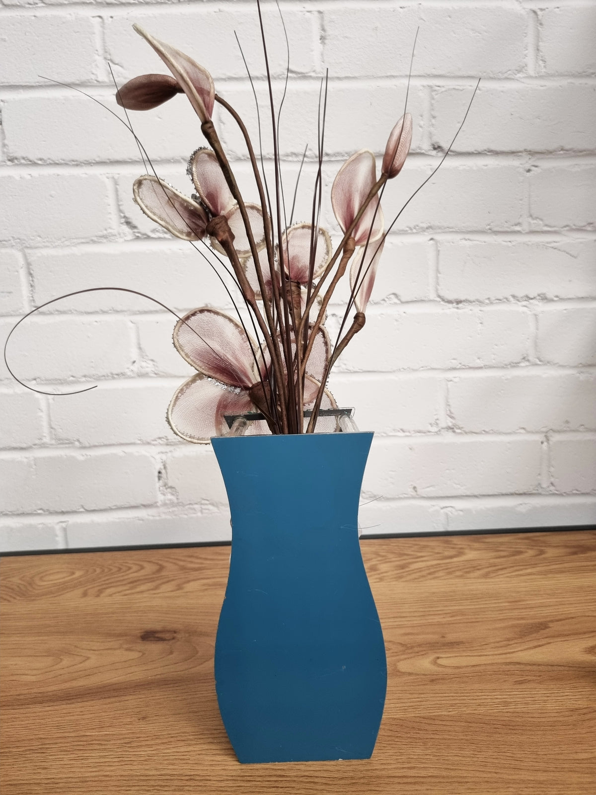 A blue ceramic vase filled with dried decorative branches and pale pink flower buds, set on a wooden floor against a white brick wall.