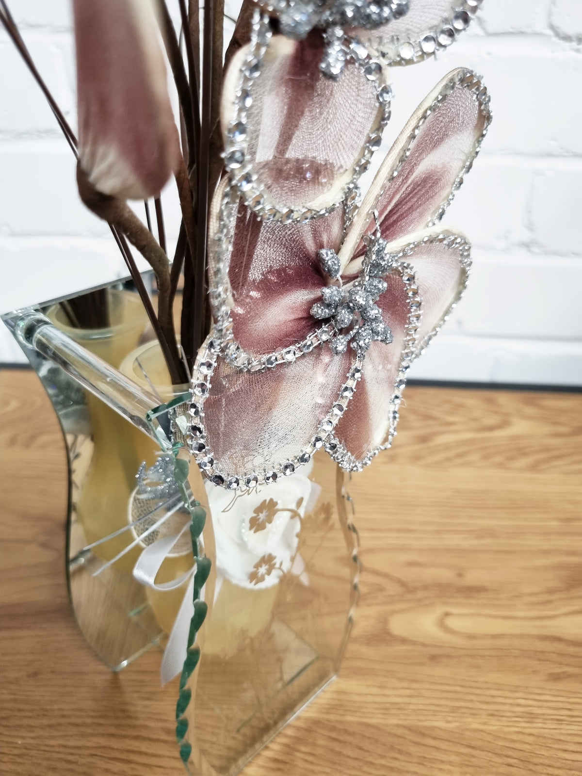A decorative floral arrangement in a clear glass vase on a wooden surface against a white brick wall. The arrangement features large dusty pink mesh flowers with silver rhinestone trim and brown dried stems.