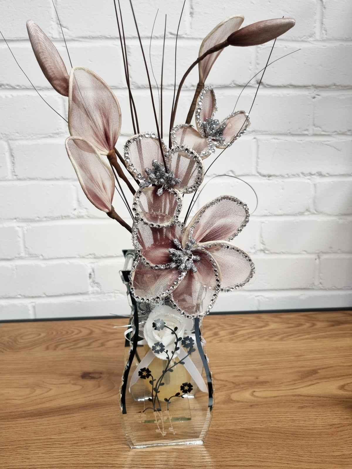A decorative floral arrangement featuring large dusty pink satin petals with silver rhinestone trim, set in a clear glass vase on a wooden tabletop against a white brick wall.