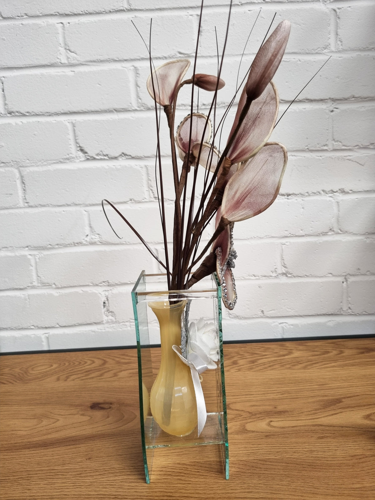 A decorative floral arrangement with pale pink calla-lily-like blooms and long brown stems in a clear rectangular glass vase with a yellow center, placed on a wooden surface against a white brick wall.