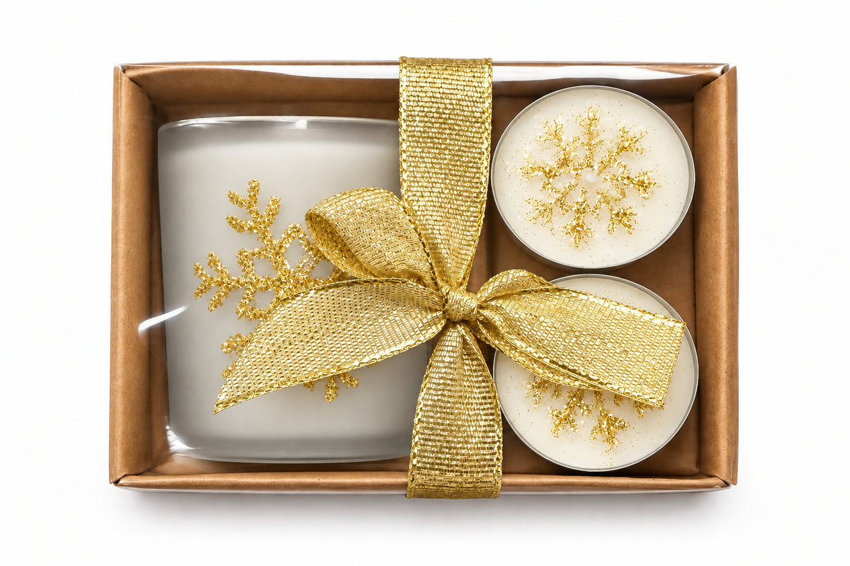 A gift set in a brown box featuring two round white candles with gold glitter, a grey glass holder in a clear sleeve, and a shiny gold ribbon bow with a gold snowflake decoration, all photographed from above on a gray textured surface.