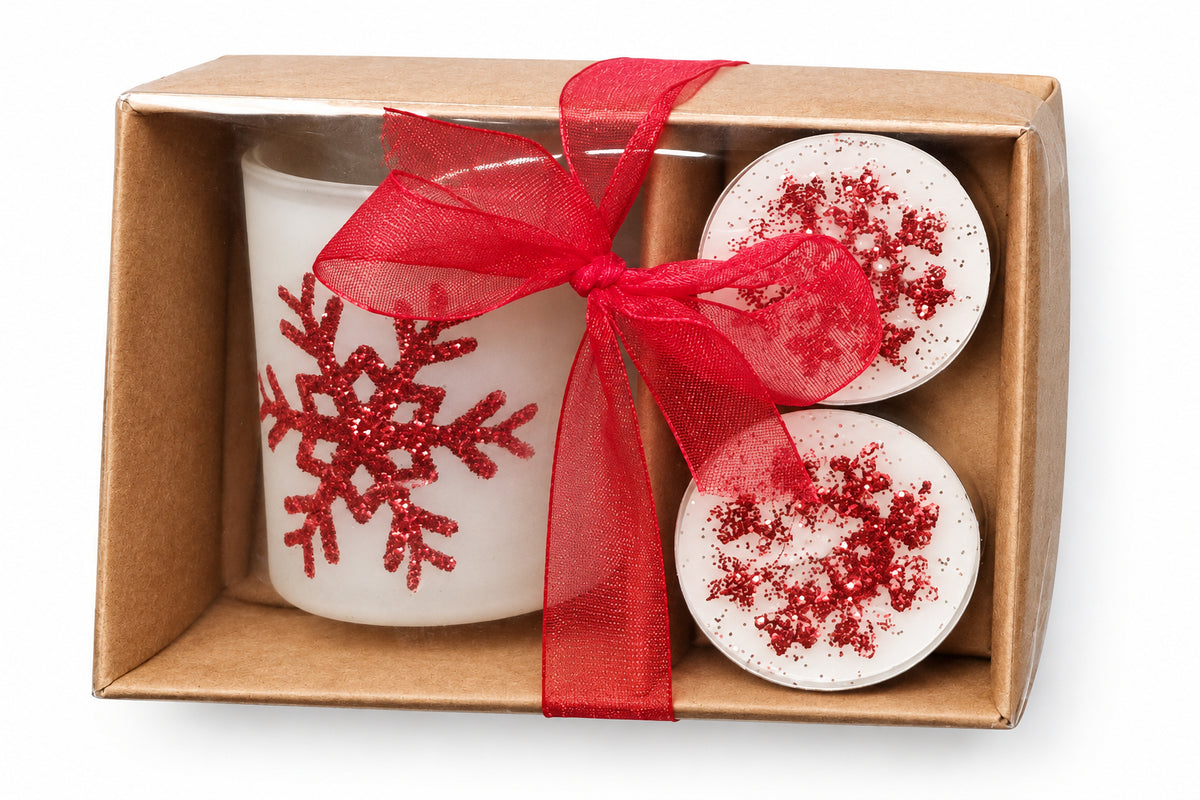 A boxed holiday gift set containing two white tea light candles topped with red glitter snowflakes and a grey glass holder with a red glitter snowflake design, tied together with a red sheer ribbon in a brown cardboard box.