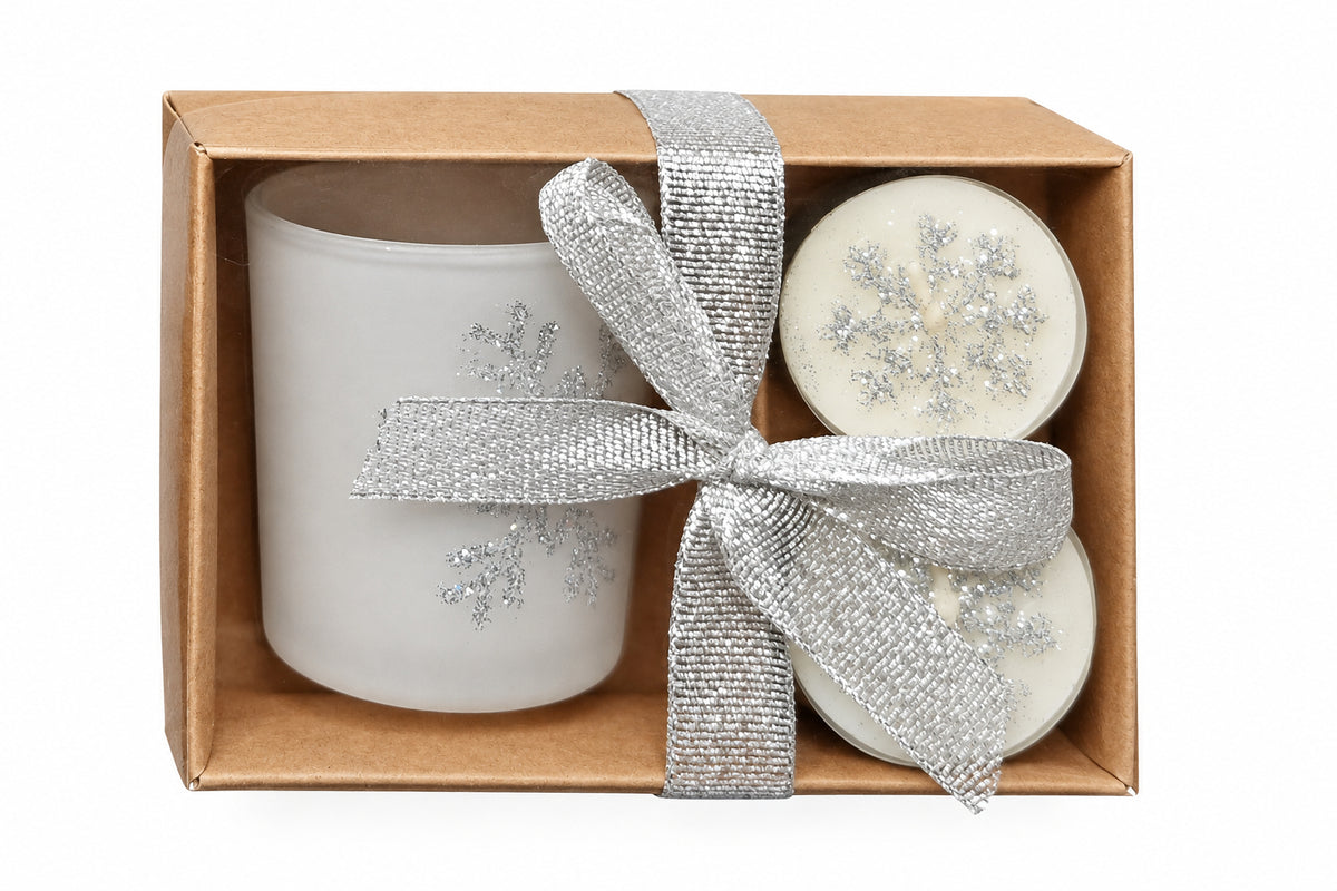 A gift set in a brown kraft box featuring a frosted glass votive, two small round wax melts or candles with silver glitter, and a silver glitter ribbon bow, set against a white background