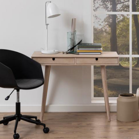 A light natural wood writing desk with two drawers and tapered legs, styled in a home office setting with a black office chair, white desk lamp, and books.
