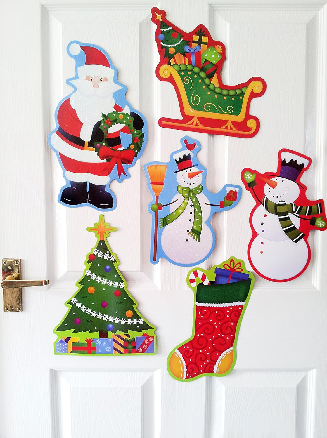 Six colorful Christmas-themed paper cutout decorations displayed on a white paneled door, including Santa Claus, a sleigh, a Christmas tree, a snowman, a stocking, and another snowman.