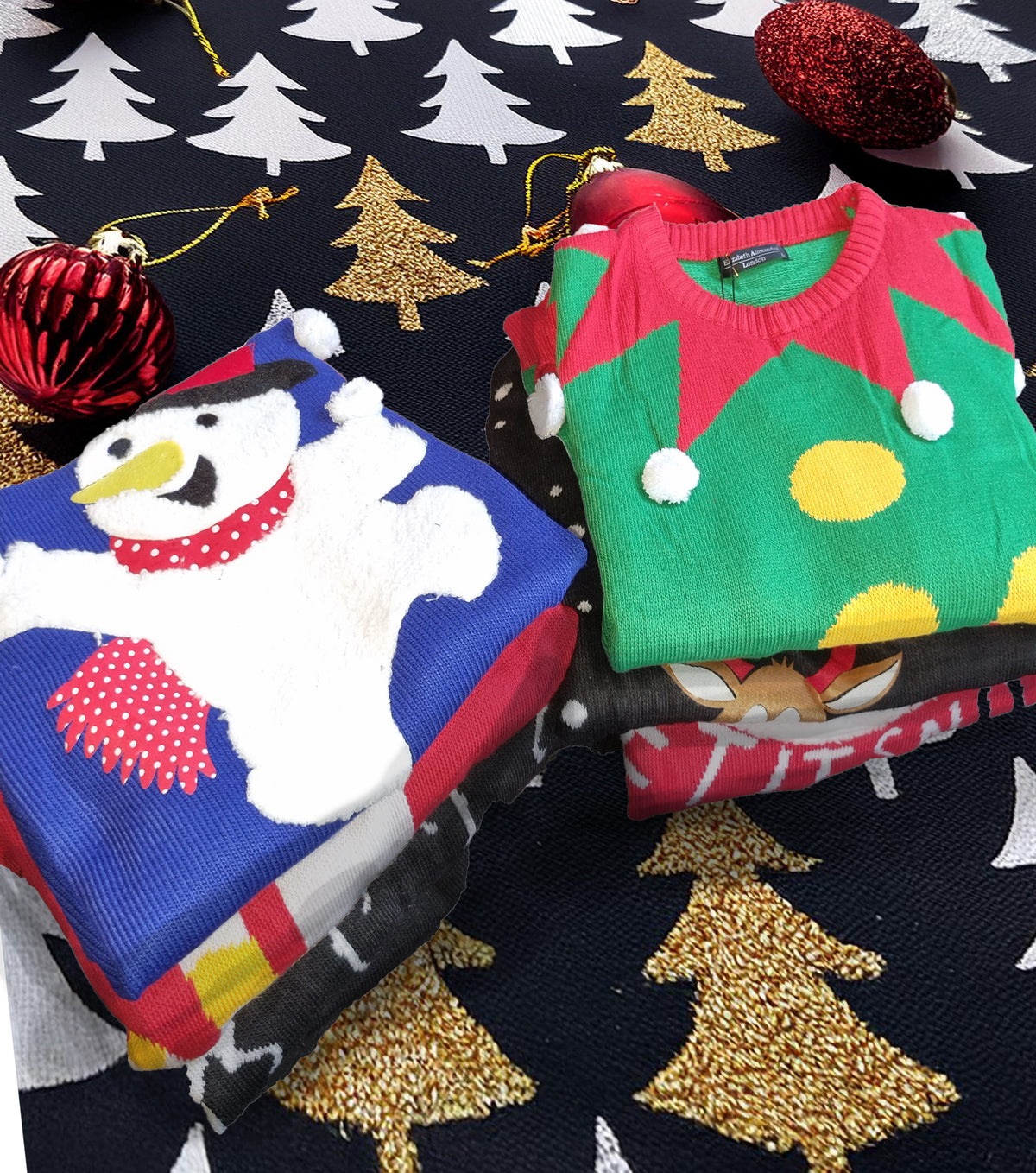 Two folded Christmas-themed knit sweaters displayed on a dark patterned fabric background with gold and white tree motifs and red ornaments. The left sweater is blue with a white snowman and red accents; the right sweater is green with red elf hat details and yellow ornaments.