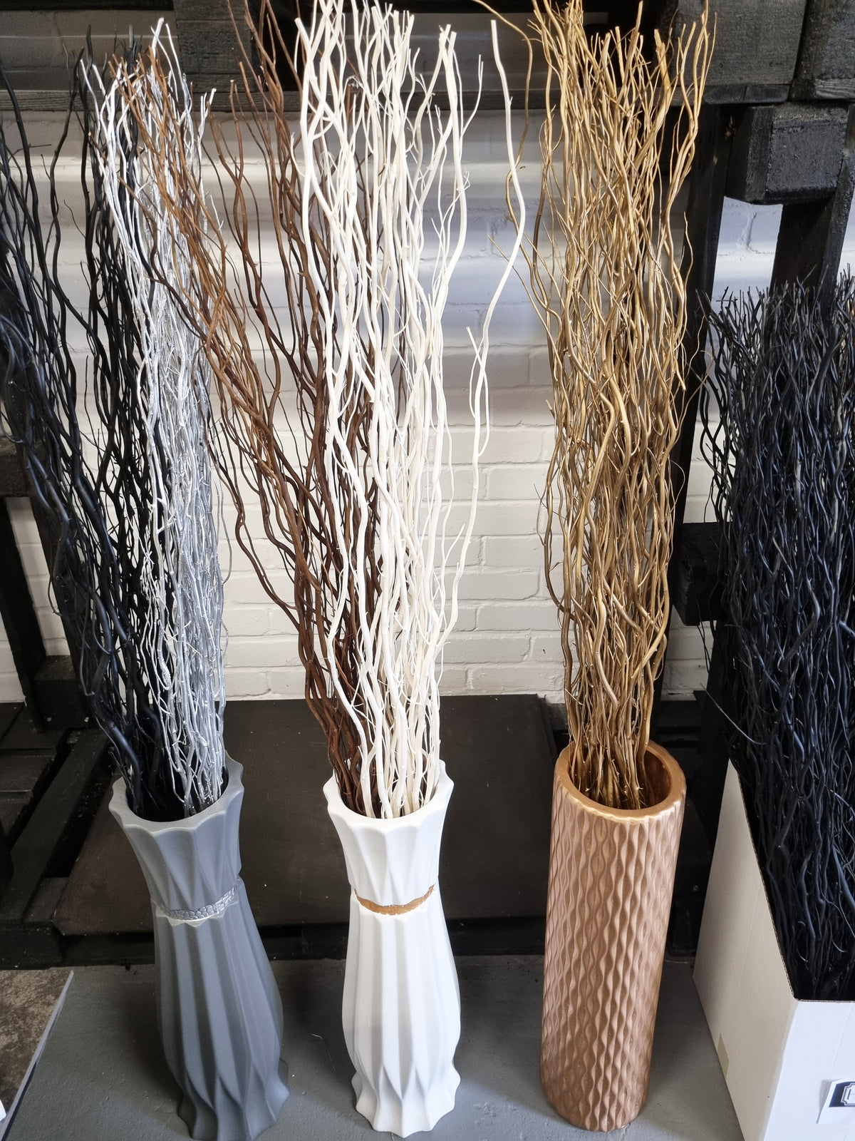 Three decorative tall dried-look branches arrangements in different colored vases, displayed indoors against a white brick wall and dark shelving.