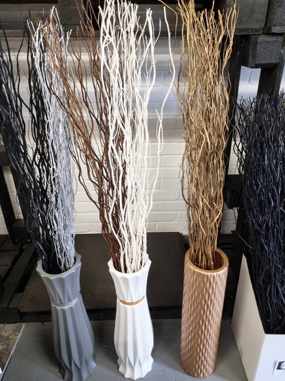 Three decorative tall dried-look branches arrangements in different colored vases, displayed indoors against a white brick wall and dark shelving.