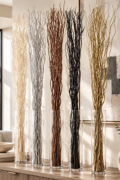 Five tall clear glass vases filled with dried decorative branches in cream, gray, brown, black, and gold tones, arranged on a wooden console in a styled interior room.