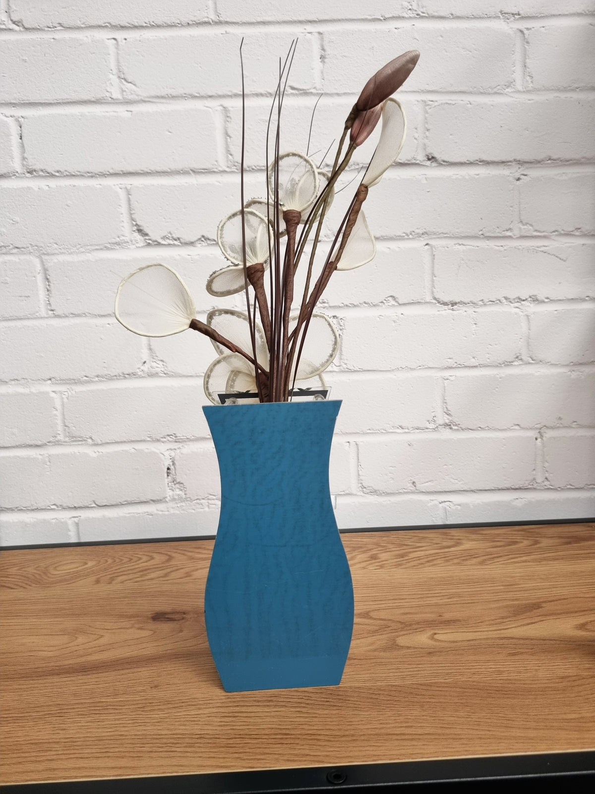 A blue textured ceramic vase filled with dried decorative grasses and seed pods, set on a wooden surface against a white brick wall background.