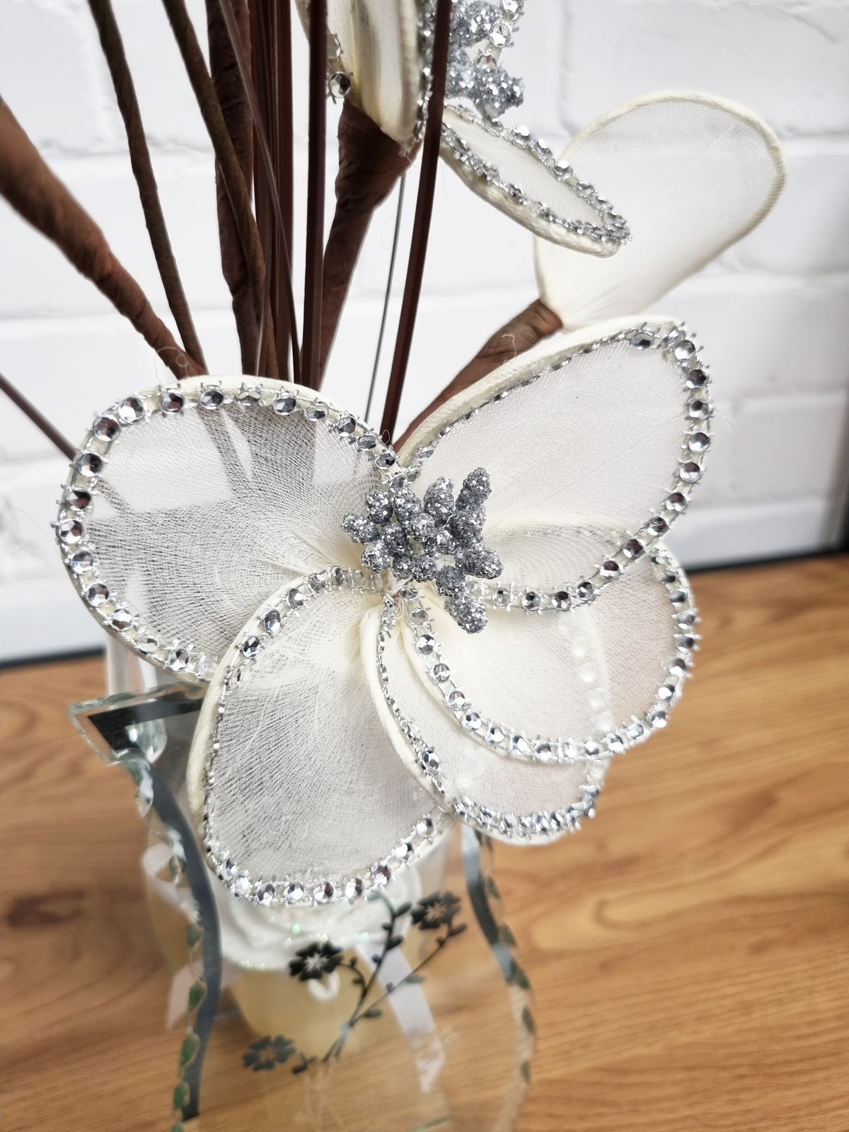 A decorative white mesh butterfly ornament embellished with silver rhinestones and a central silver glittery flower, displayed in a glass vase with dried brown stems on a wooden surface.