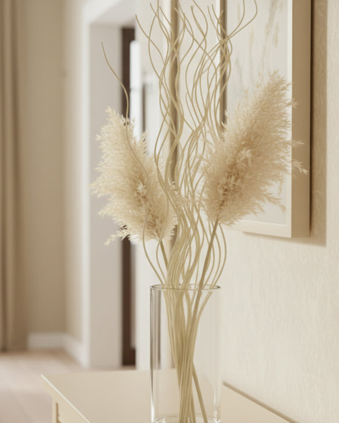 A clear glass vase filled with dried pampas grass and tall, slender decorative branches, placed on a light wood console table in a softly lit interior hallway.