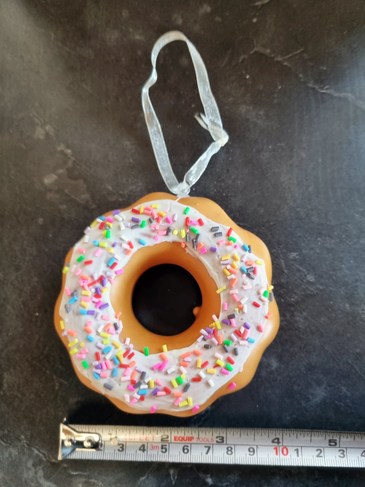 A novelty donut-shaped ornament with white frosting and colorful sprinkles, hanging from a clear ribbon, shown on a dark surface with a measuring tape below for scale.