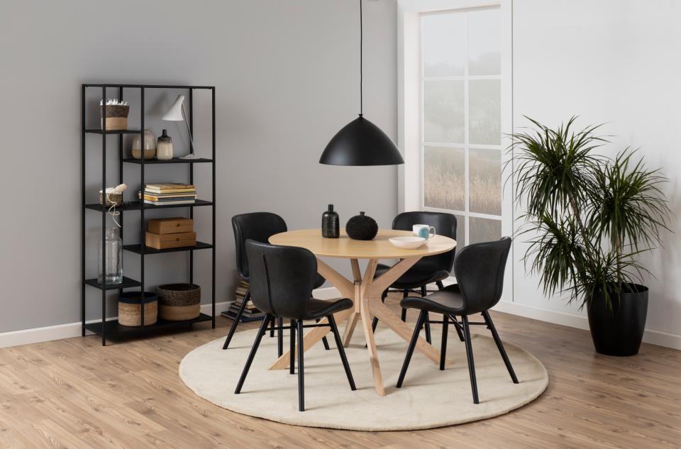 A modern dining room setup featuring a round light wood table with a star-shaped wooden base, surrounded by four black chairs. The set is styled on a round beige rug under a black dome pendant light, set against a gray wall with a black shelving unit and a potted plant.