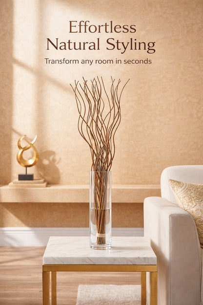 A clear cylindrical glass vase filled with dried brown branches, displayed on a marble-top side table in a styled living room. A cream sofa with a patterned pillow is visible on the right, and the wall above reads 'Effortless Natural Styling' with smaller text 'Transform any room in seconds'.